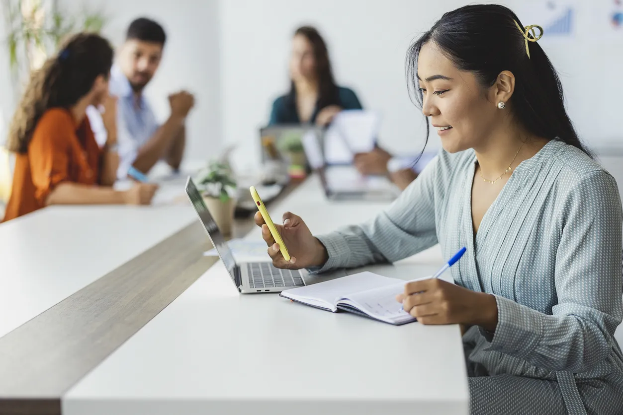 Woman at a shared counter with laptop, phone, pen, and notebook