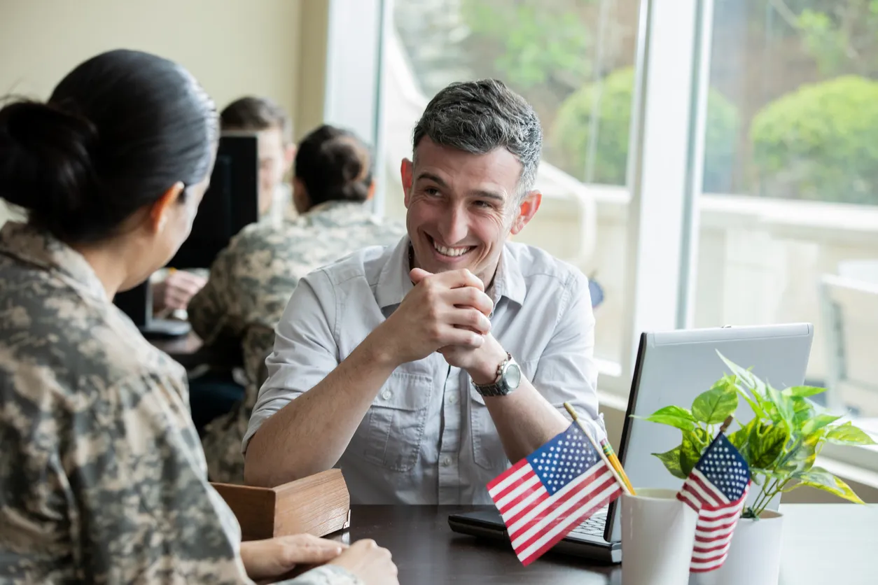 Woman in a military uniform and a man at a desk with a laptop and a cup with small American flags