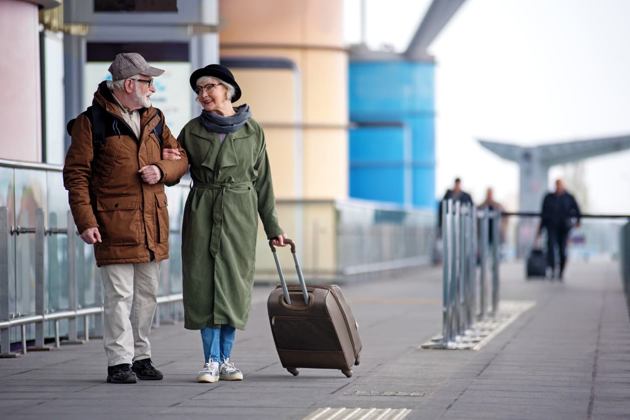 Full length of encouraged calm retired man and woman are talking to each other while standing outdoors. They are carrying a suitcase while looking at each other with love.
