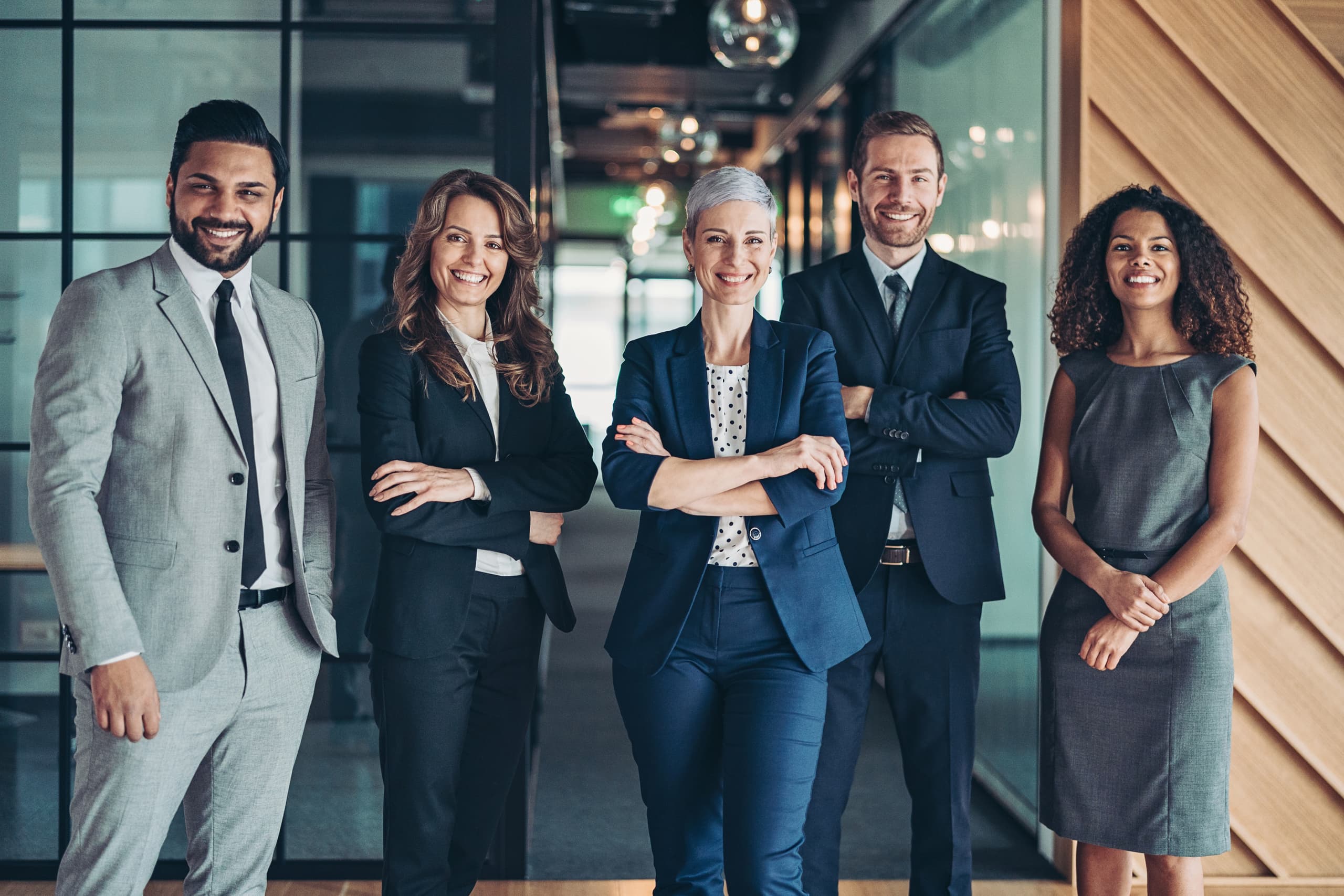 Five professionally dressed people smiling in an office hallway